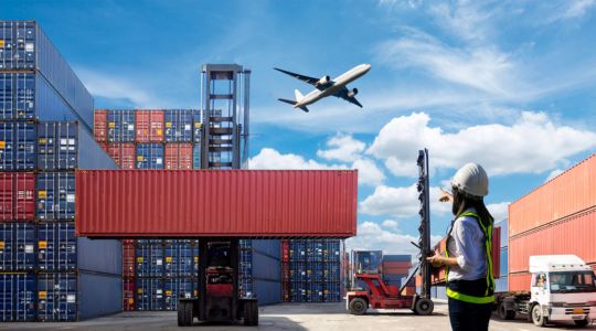 An airplane flying above shipping containers, trucks, and a worker wearing a safety vest on the ground.