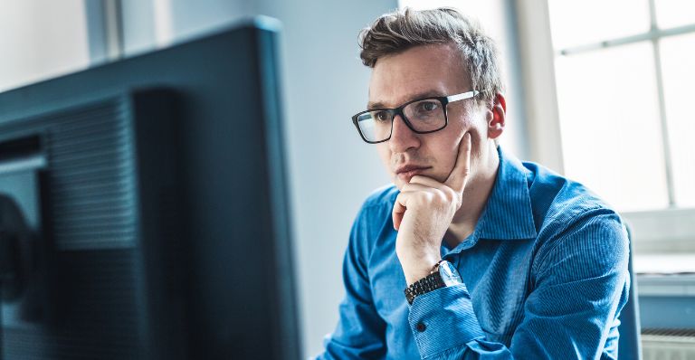 A young businessman wearing eyeglasses is seated at a desk in an office, intently focused on his computer monitor.