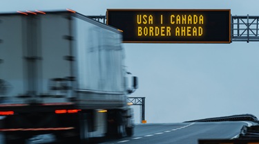 Transport truck passes under USA-Canada Border ahead sign on highway.